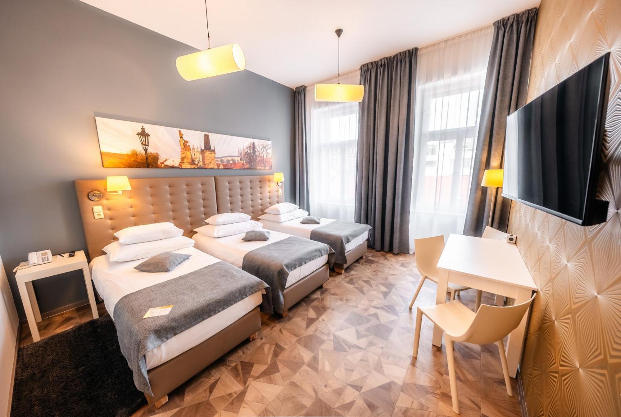 Grandium Hotel Prag Dış mekan fotoğraf The photo shows a modern hotel room featuring three single beds, each with white bedding and gray throws. The walls are painted gray, and artwork depicting a cityscape is displayed above the headboard. There are two stylish pendant lights hanging fro