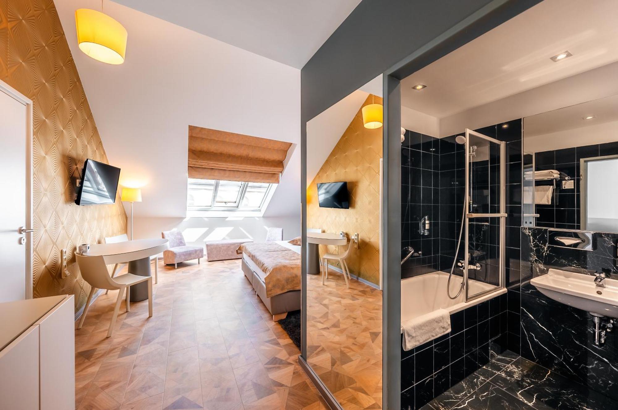 Grandium Hotel Prag Dış mekan fotoğraf The photo shows an interior of a hotel room featuring a modern design. On one side, there are two beds with white linens, a desk, and a television mounted on the wall. The room has sloped ceilings and large windows that let in natural light. 

Adjace