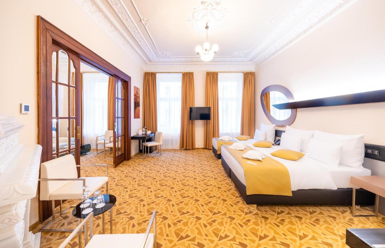Grandium Hotel Prag Dış mekan fotoğraf The photo shows a spacious hotel room with an elegant design. There are two large beds with white bedding, accented with yellow pillows. The room has tall windows with flowing curtains that allow natural light to brighten the space. A flat-screen tel